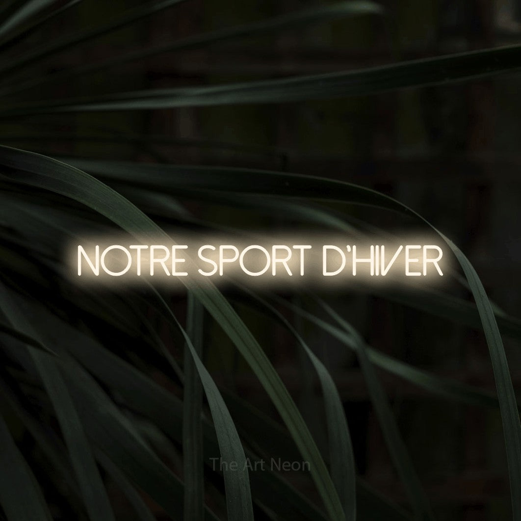 “Notre sport d'hiver” LED neon sign in warm white, with thin sans-serif lettering, shown against a close-up of dark foliage.
