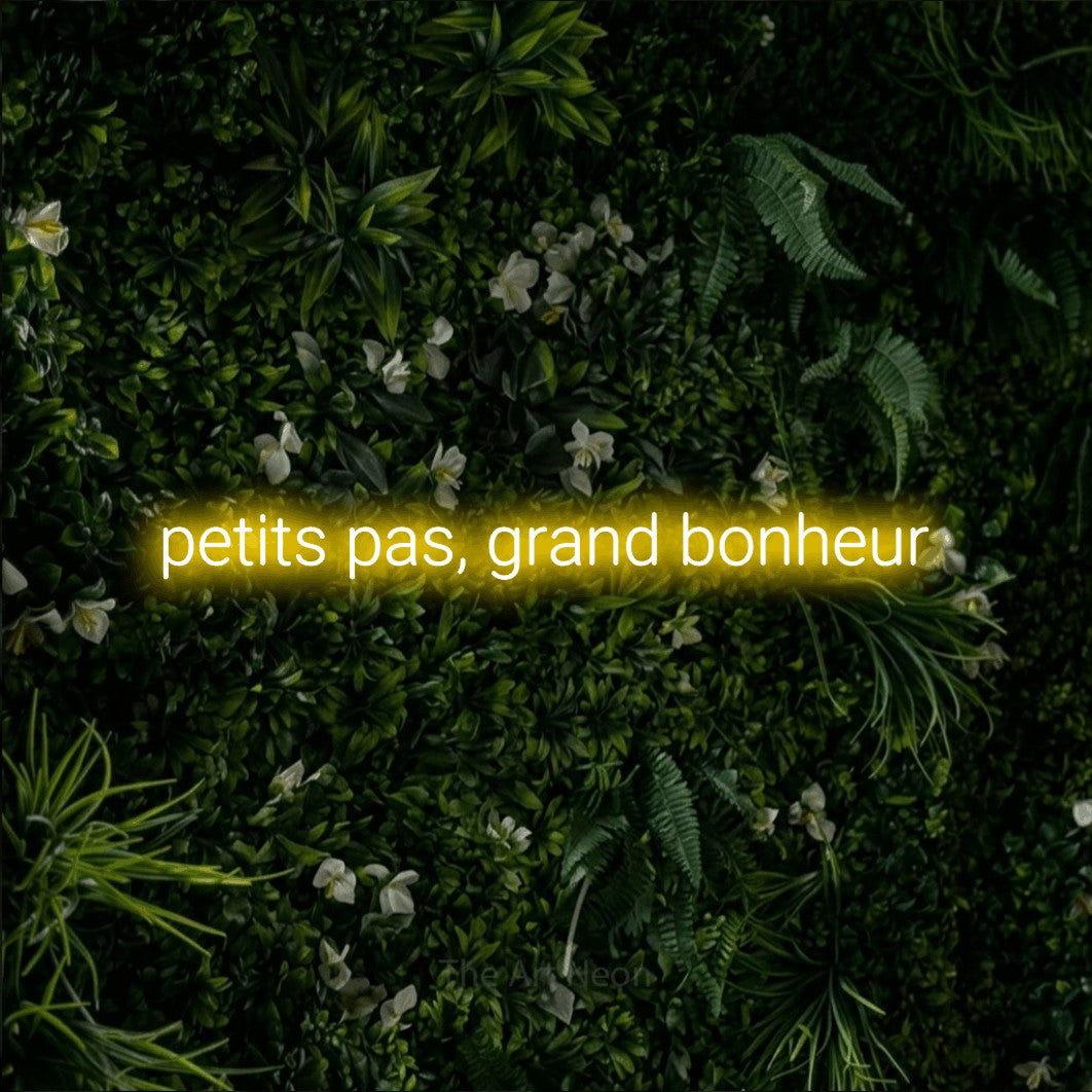 “Petits pas, grand bonheur” LED neon sign in yellow, with rounded sans-serif lettering, shown against a dense green plant wall.
