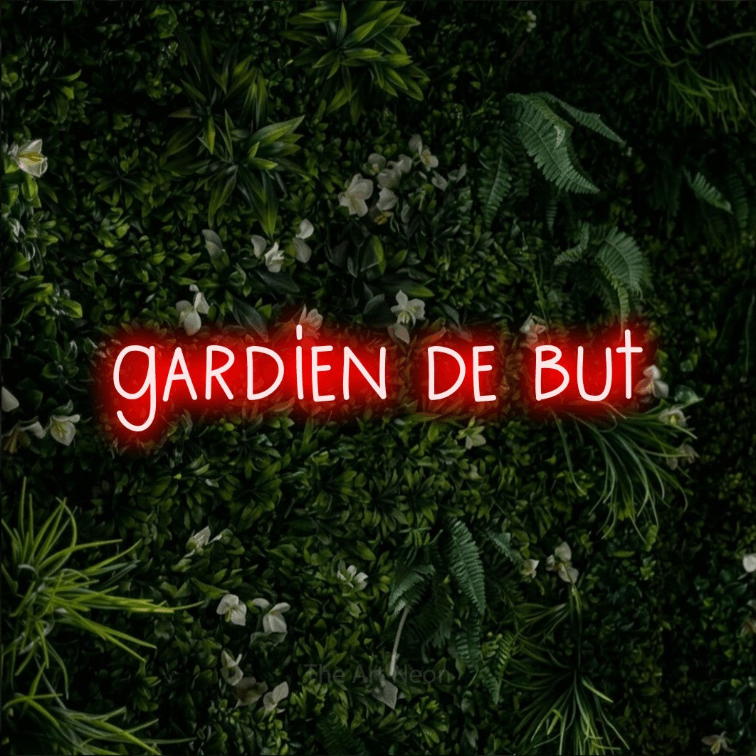 “Gardien de but” LED neon sign in red, with narrow hand-drawn lettering, shown against a dense green plant wall.