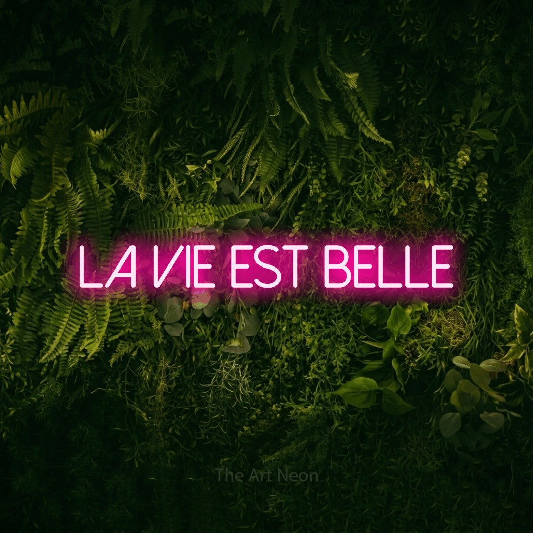 “La vie est belle” LED neon sign in hot pink, with thin sans-serif lettering, shown against a dark moss and forest backdrop.