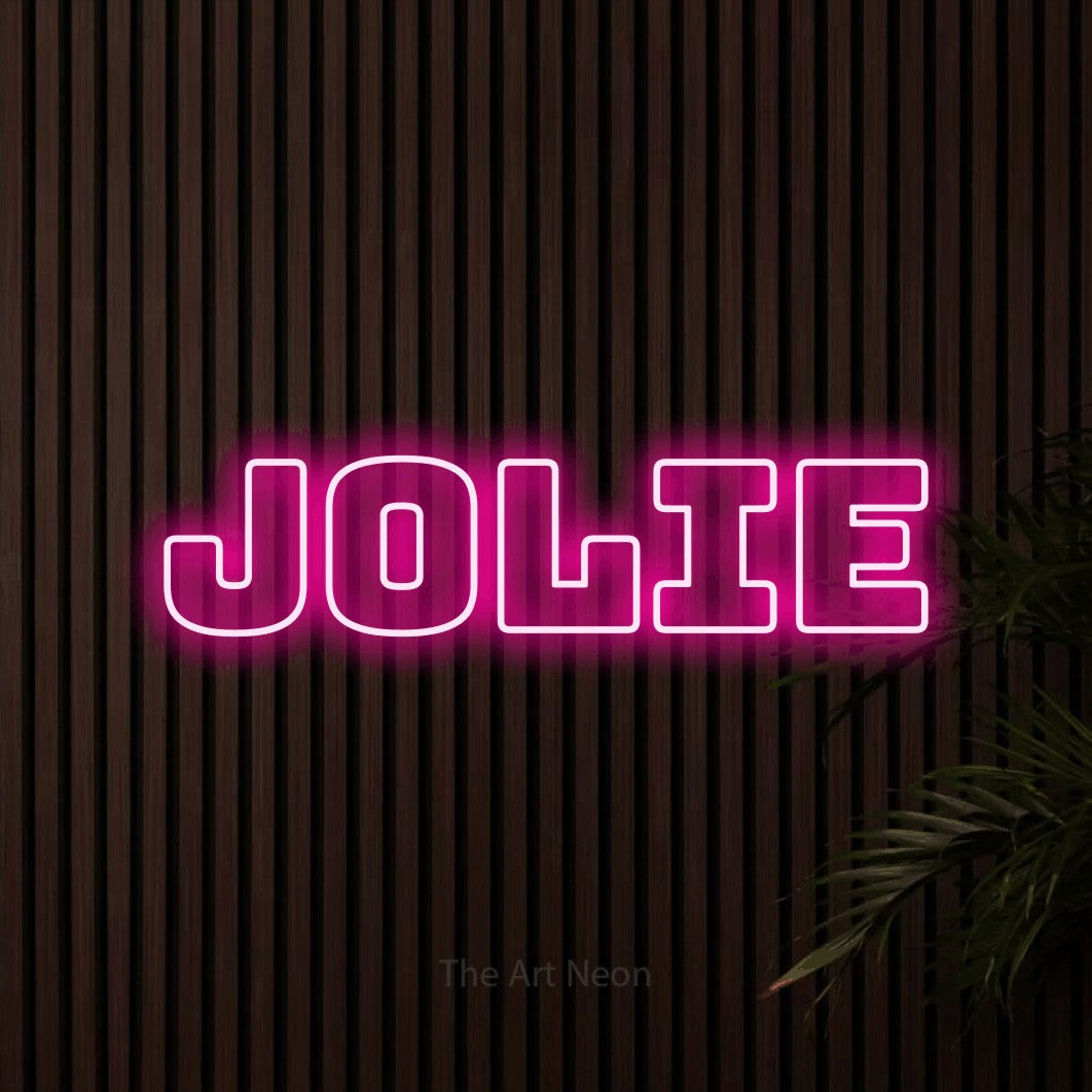 Décoration murale en néon LED « Jolie » en rose vif, en typographie contour épaisse, installée sur un mur de lattes en bois foncé, pour un salon de beauté.