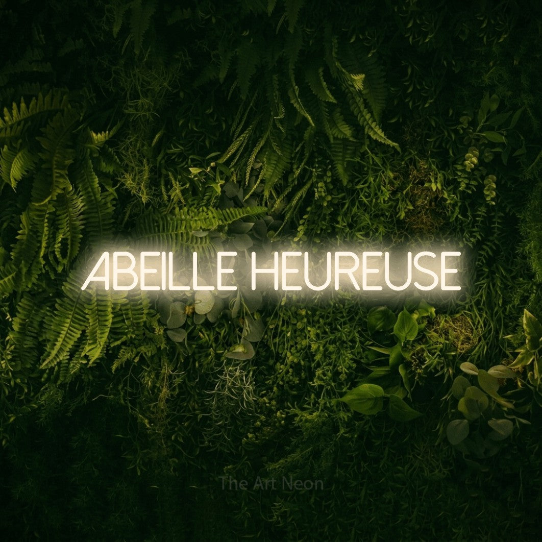 “Abeille heureuse” LED neon sign in warm white, with thin sans-serif lettering, shown against a dark moss and forest backdrop.