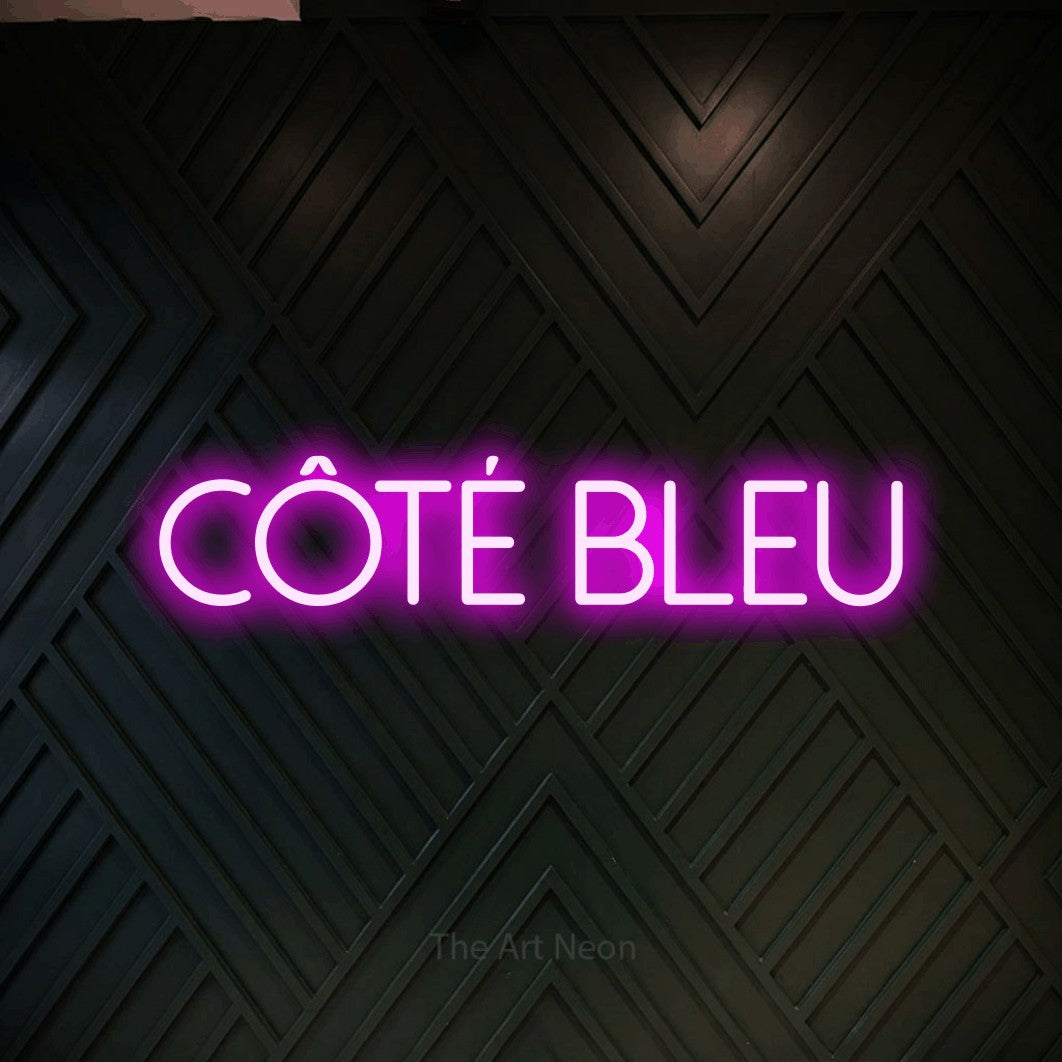 “Côté bleu” LED neon sign in pink, with thin sans-serif lettering, shown against a black chevron-patterned wood wall.