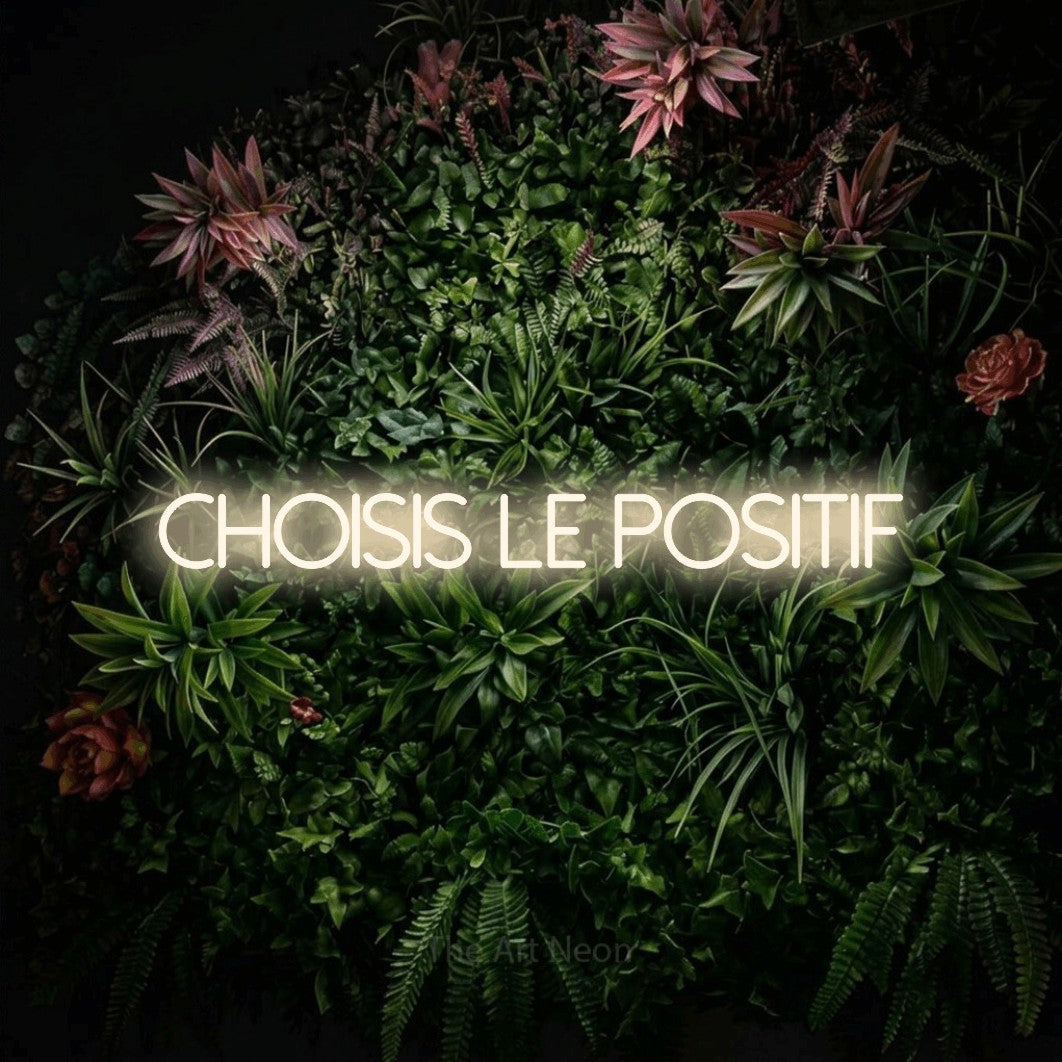 “Choisis le positif” LED neon sign in warm white, with thin sans-serif lettering, shown against a flowering plant wall.
