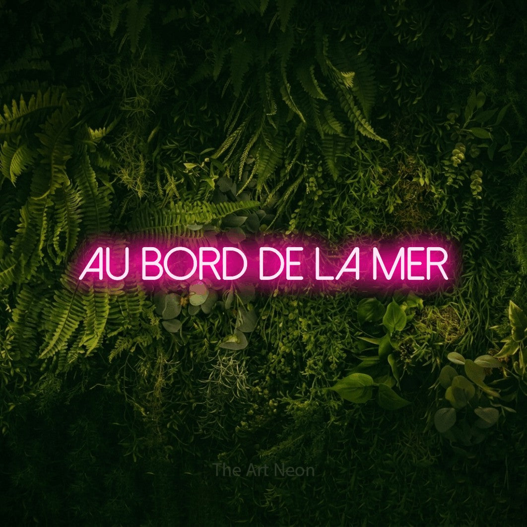 “Au bord de la mer” LED neon sign in hot pink, with thin sans-serif lettering, shown against a dark moss and forest backdrop.