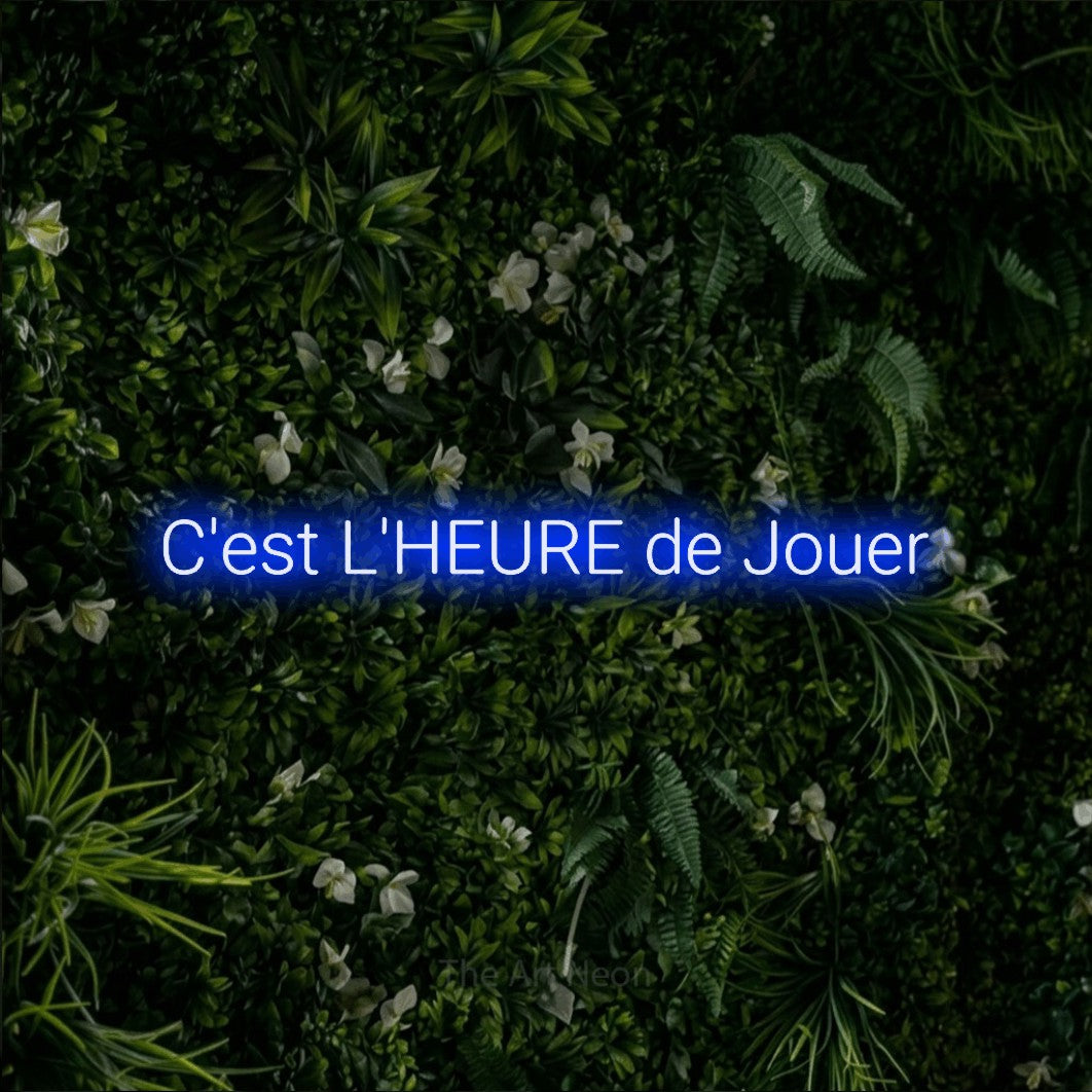 “C'est l'heure de jouer” LED neon sign in blue, with rounded sans-serif lettering, shown against a dense green plant wall.