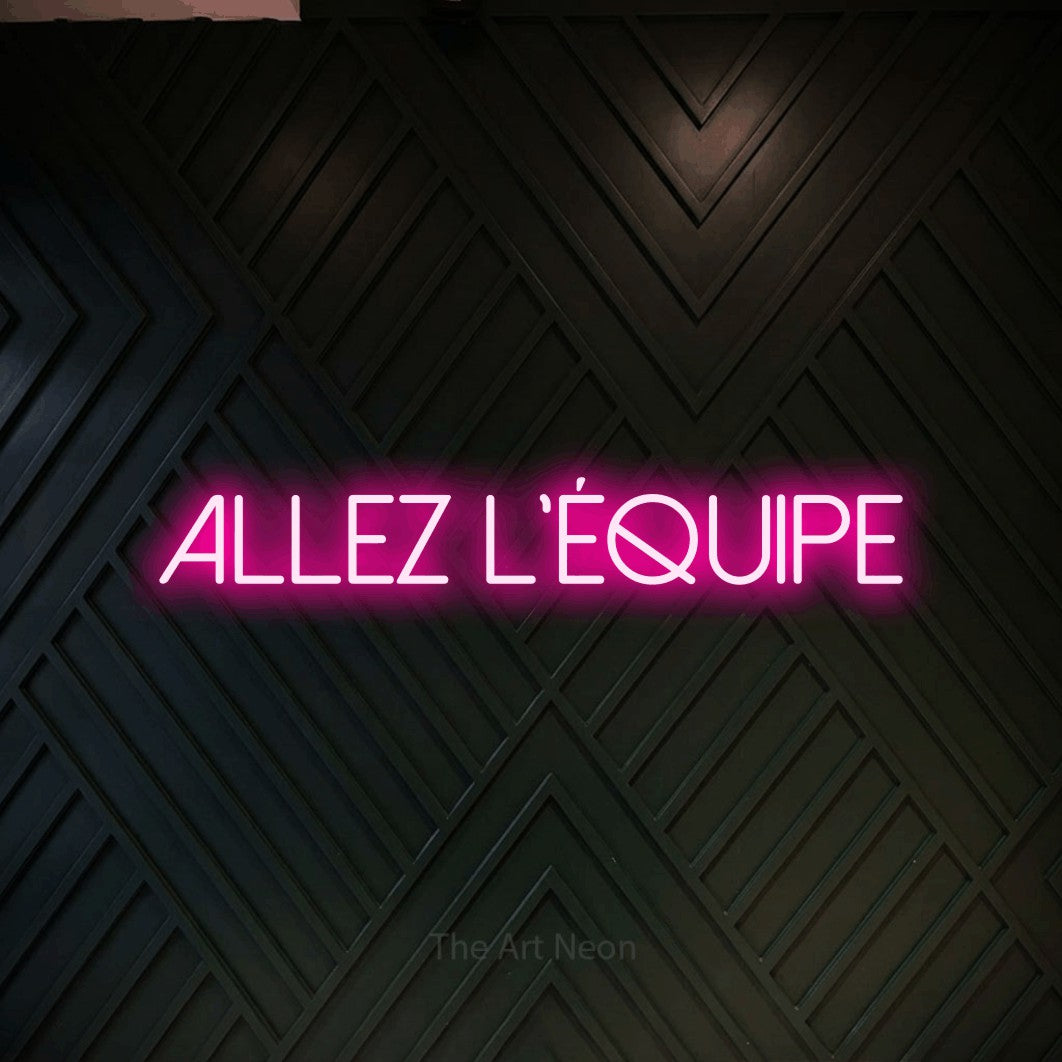 “Allez l'équipe” LED neon sign in hot pink, with thin sans-serif lettering, shown against a black chevron-patterned wood wall.