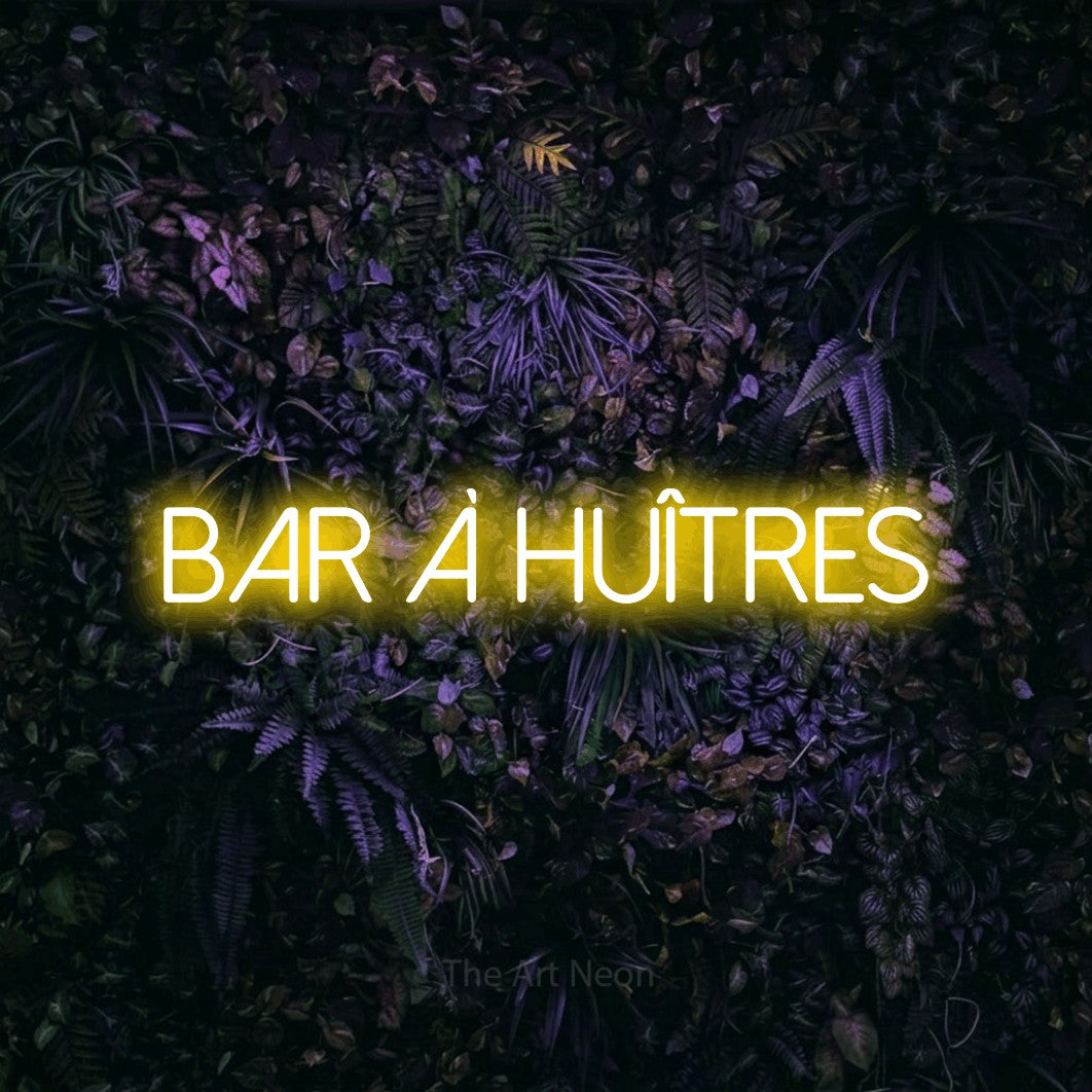 “Bar à huîtres” LED neon sign in yellow, with thin sans-serif lettering, shown against a dark wall with purple flowers.