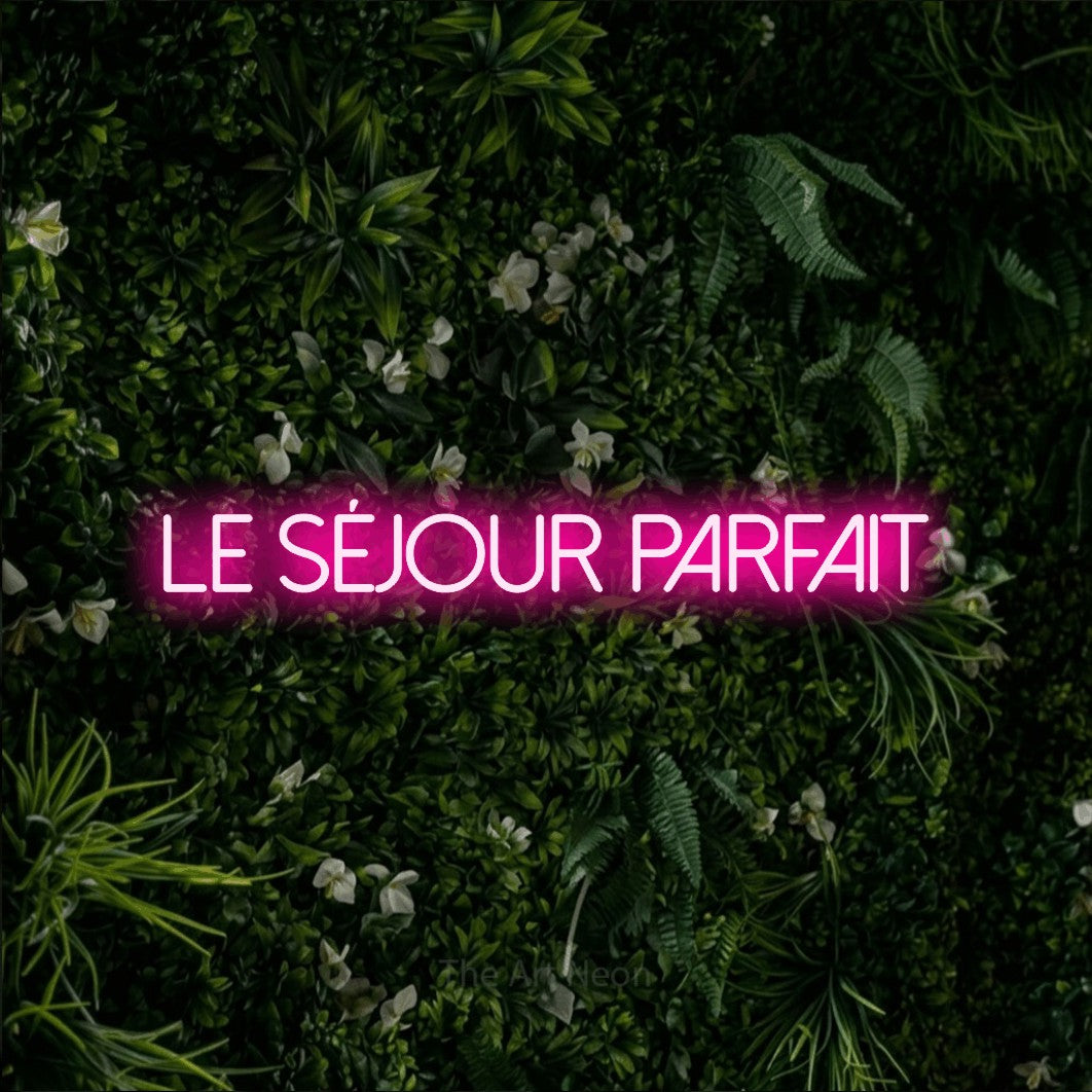 “Le séjour parfait” LED neon sign in hot pink, with thin sans-serif lettering, shown against a dense green plant wall.