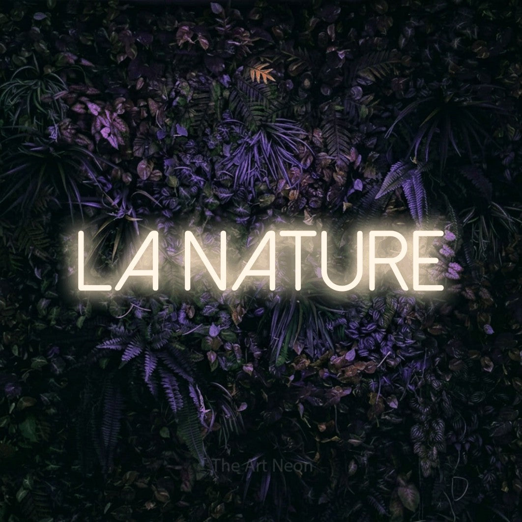 “La nature” LED neon sign in warm white, with thin sans-serif lettering, shown against a dark wall with purple flowers.