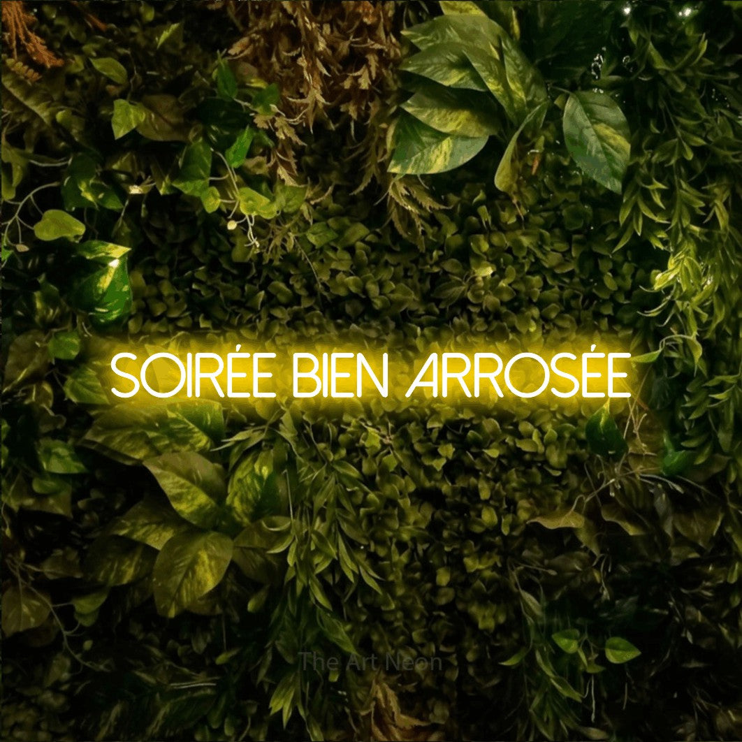 “Soirée bien arrosée” LED neon sign in yellow, with thin sans-serif lettering, shown against bright green and yellow foliage.