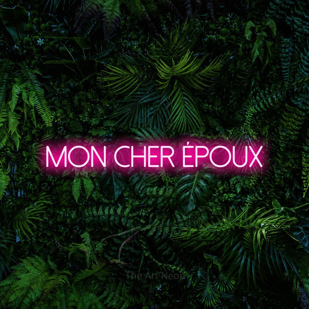 “Mon cher époux” LED neon sign in hot pink, with thin sans-serif lettering, shown against dense tropical foliage.
