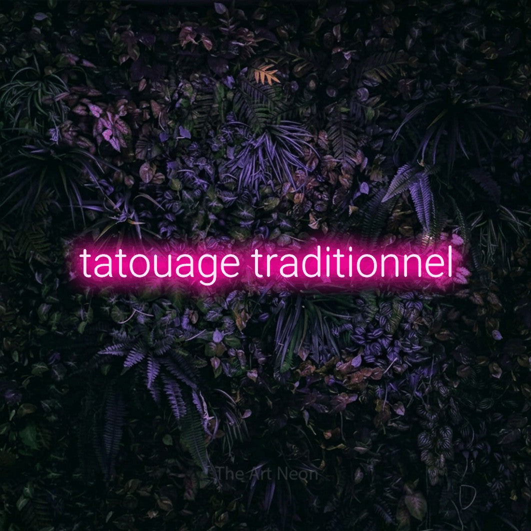 “Tatouage traditionnel” LED neon sign in hot pink, with rounded sans-serif lettering, shown against a dark wall with purple flowers.