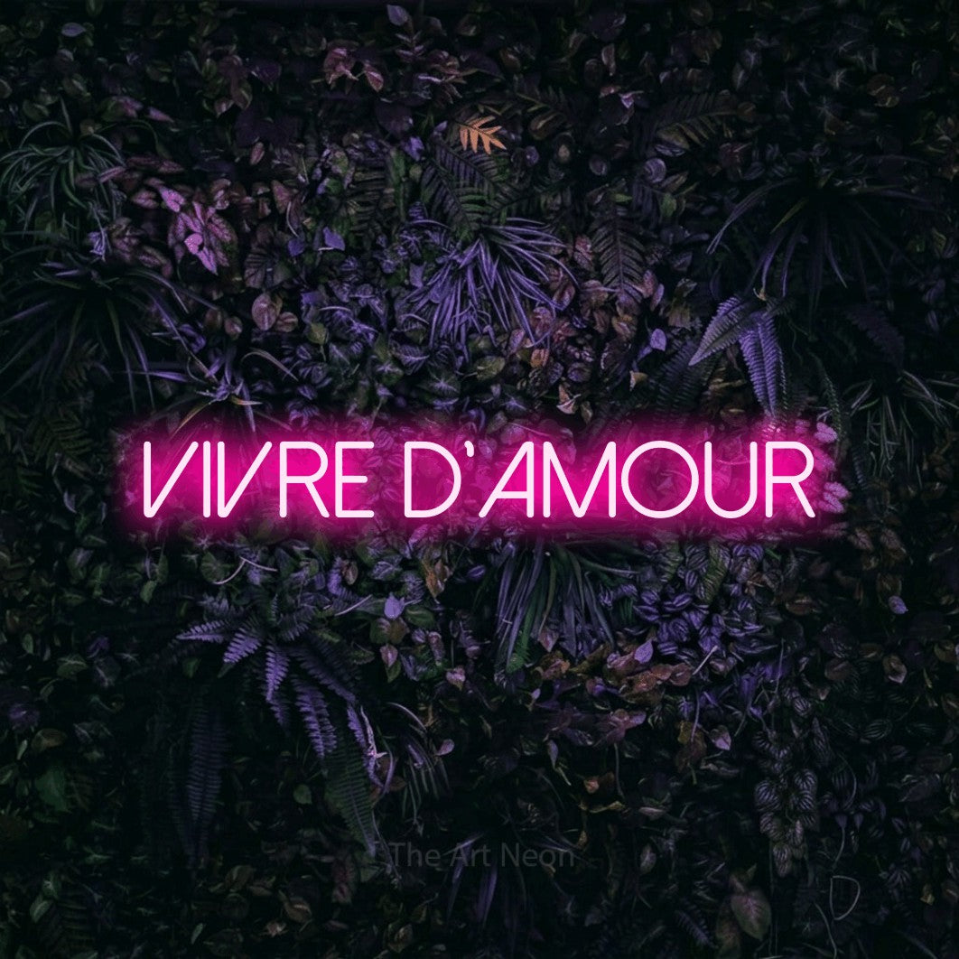 “Vivre d'amour” LED neon sign in hot pink, with thin sans-serif lettering, shown against a dark wall with purple flowers.
