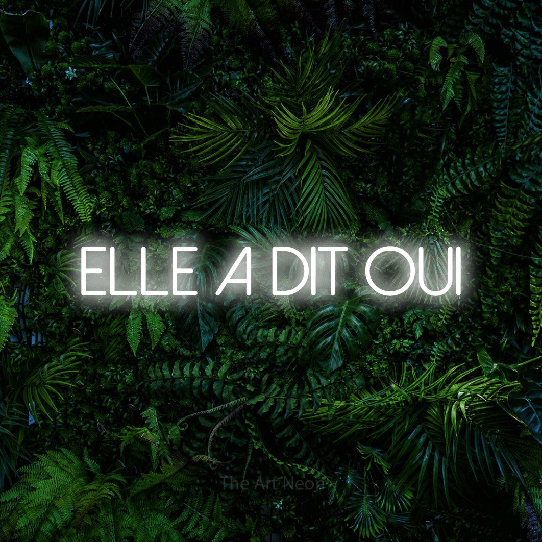 “Elle a dit oui” LED neon sign in cool white, with thin sans-serif lettering, shown against dense tropical foliage.