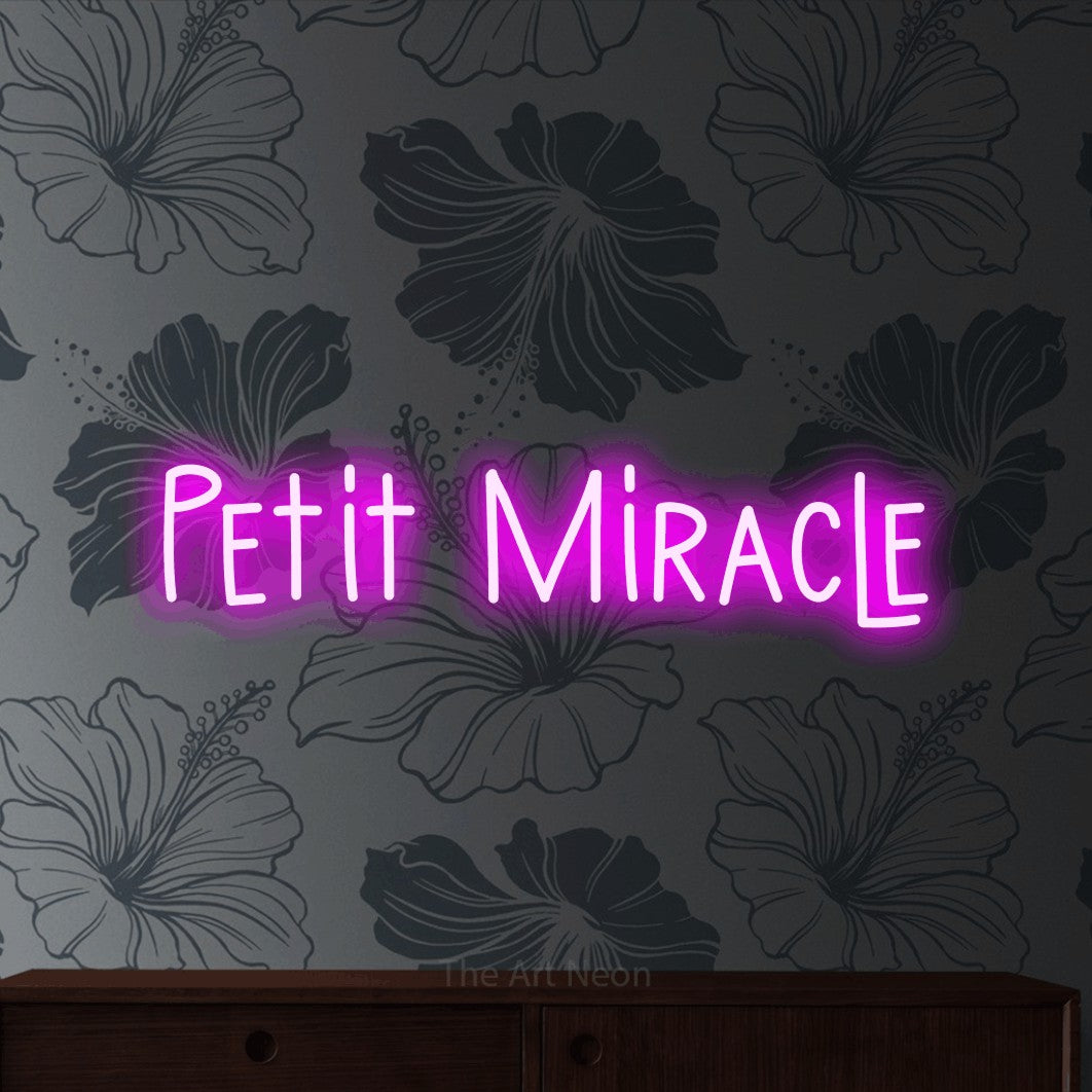 Néon LED « Petit miracle » en rose, en lettres étroites dessinées à la main, mis en scène sur un mur gris à motifs floraux, pour une chambre d’enfant.
