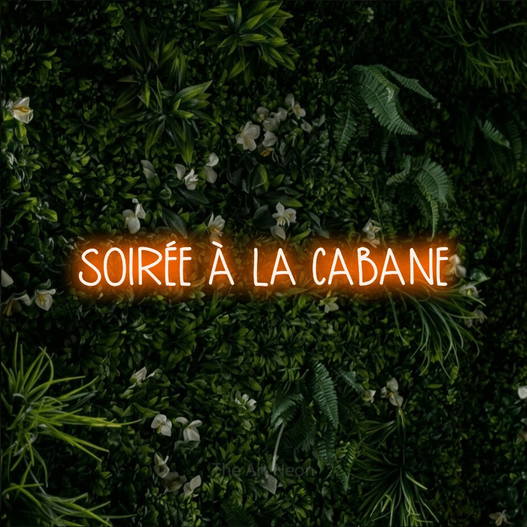 “Soirée à la cabane” LED neon sign in orange, with narrow hand-drawn lettering, shown against a dense green plant wall.