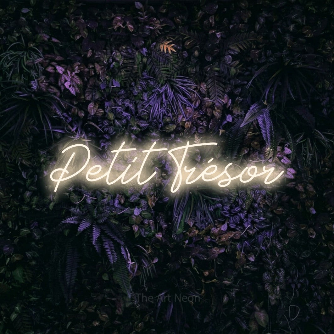 “Petit trésor” LED neon sign in warm white, with cursive lettering, shown against a dark wall with purple flowers.