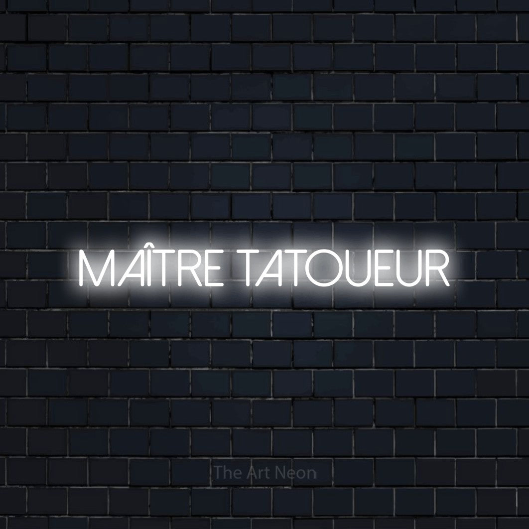 “Maître tatoueur” LED neon sign in cool white, with thin sans-serif lettering, shown against a dark navy brick wall.