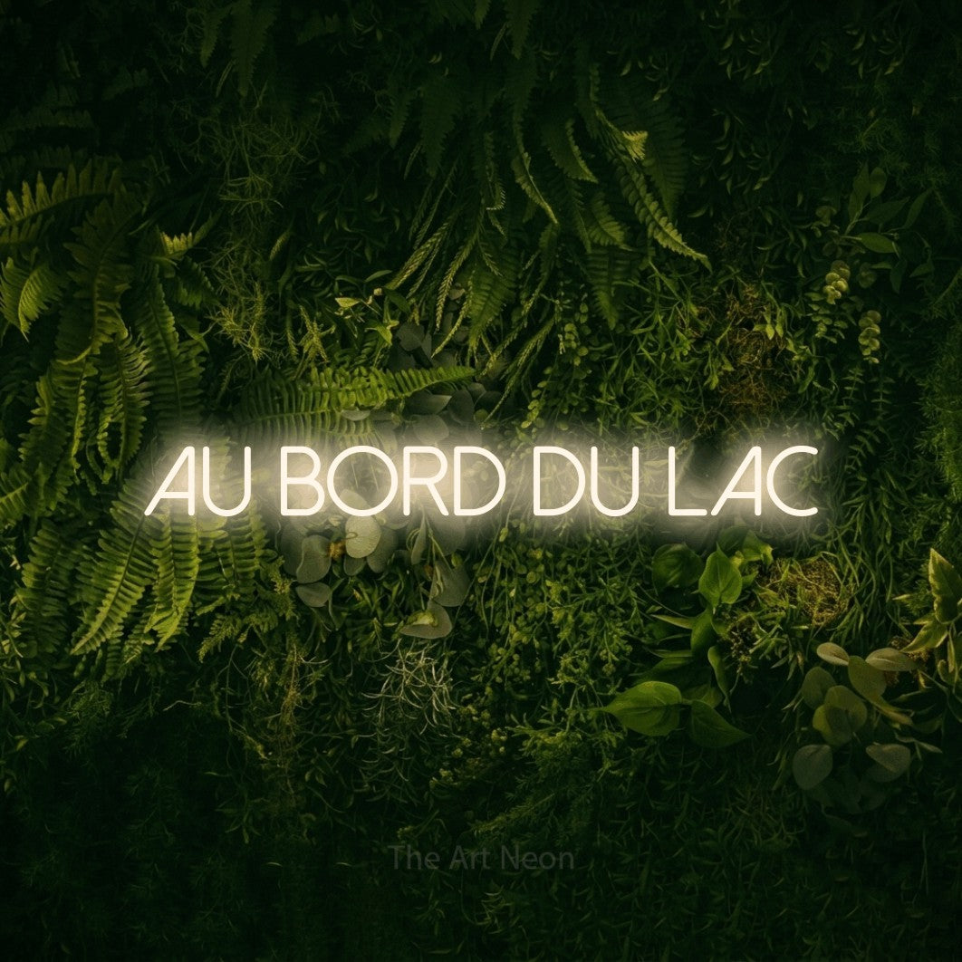 “Au bord du lac” LED neon sign in warm white, with thin sans-serif lettering, shown against a dark moss and forest backdrop.