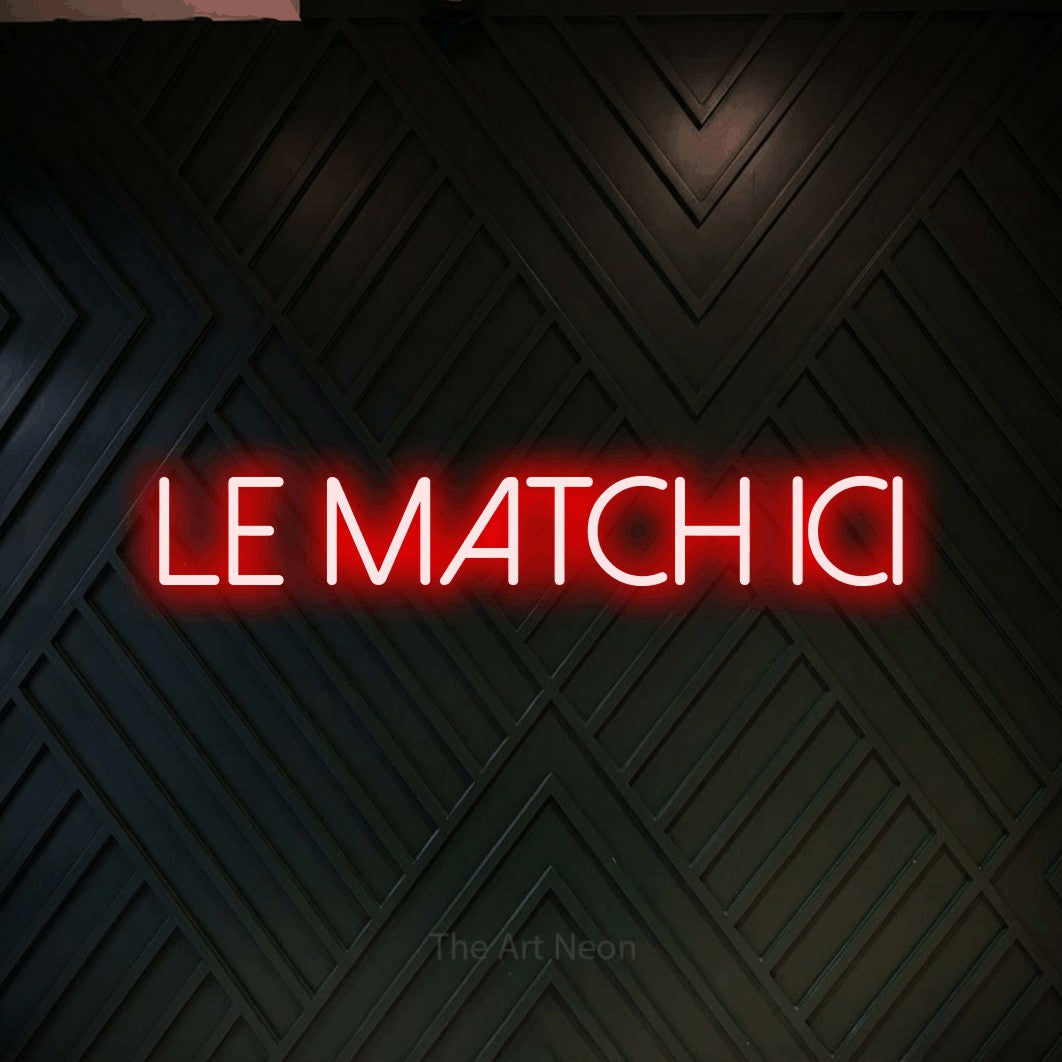 “Le match ici” LED neon sign in red, with thin sans-serif lettering, shown against a black chevron-patterned wood wall.