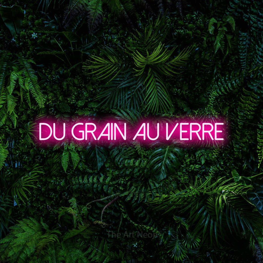 “Du grain au verre” LED neon sign in hot pink, with thin sans-serif lettering, shown against dense tropical foliage.