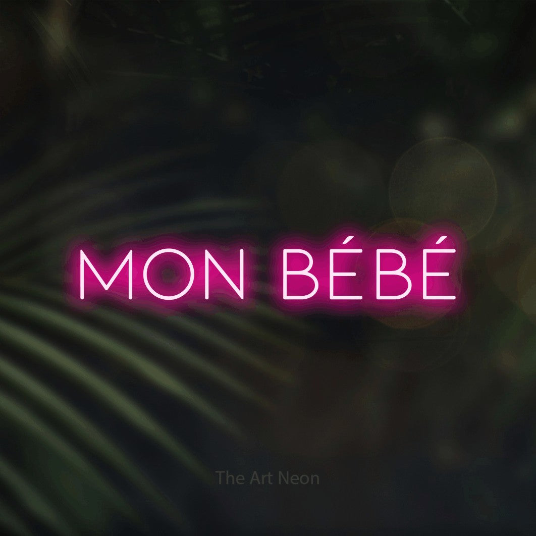 “Mon bébé” LED neon sign in hot pink, with thin, widely spaced lettering, shown against a blurred palm and bokeh-light backdrop.