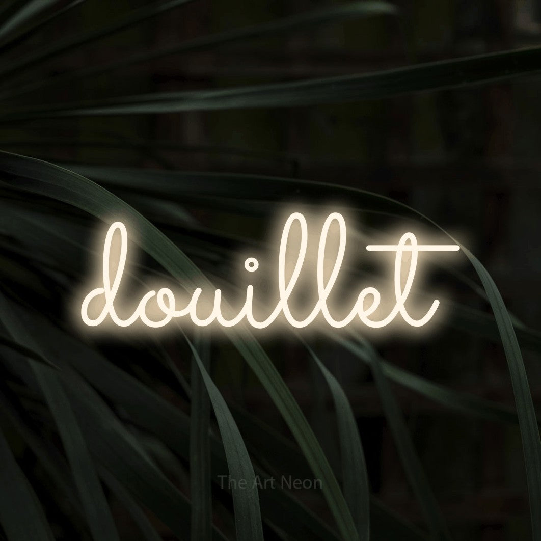 “Douillet” LED neon sign in warm white, with looped cursive lettering, shown against a close-up of dark foliage.