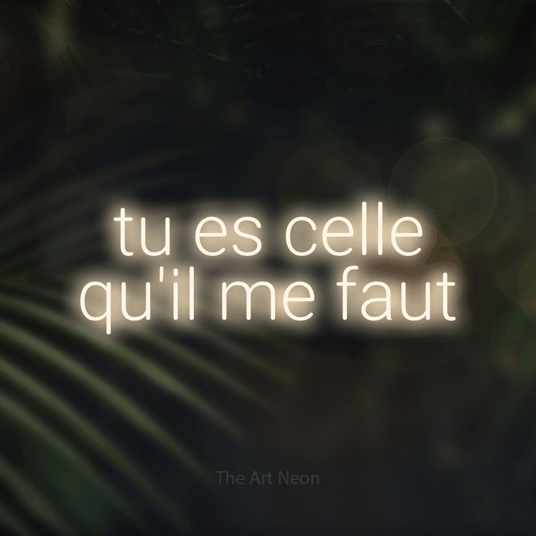 “Tu es celle qu'il me faut” LED neon sign in warm white, with rounded sans-serif lettering across 2 lines, shown against a blurred palm and bokeh-light backdrop.