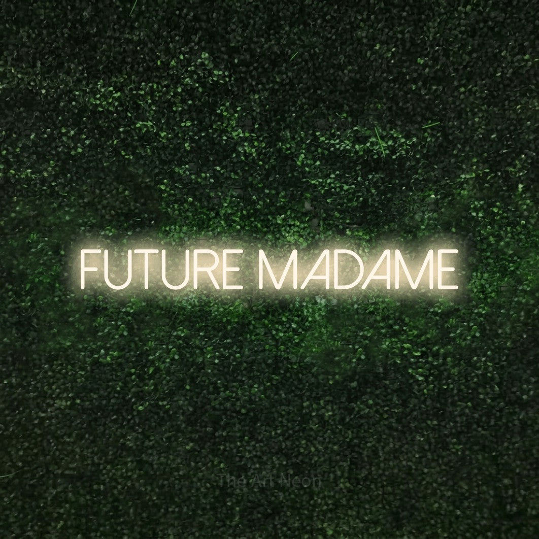 “Future madame” LED neon sign in warm white, with thin sans-serif lettering, shown against a textured green hedge.