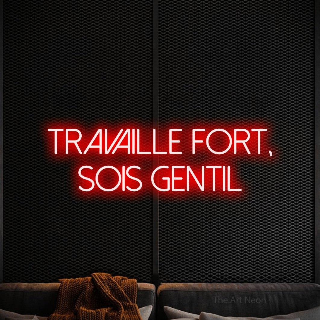 “Travaille fort, sois gentil” LED neon sign in red, with thin sans-serif lettering across 2 lines, shown against a modern dark-toned seating area.