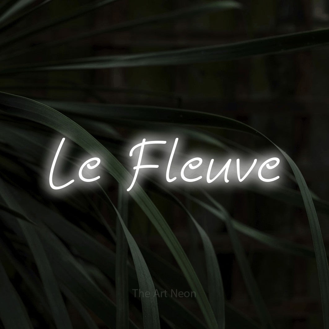 “Le fleuve” LED neon sign in cool white, with slanted handwritten lettering, shown against a close-up of dark foliage.