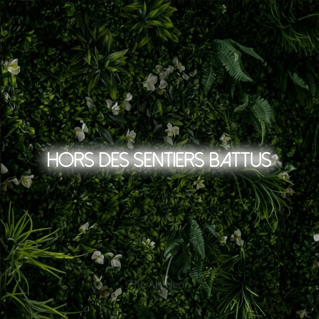 “Hors des sentiers battus” LED neon sign in cool white, with thin sans-serif lettering, shown against a dense green plant wall.