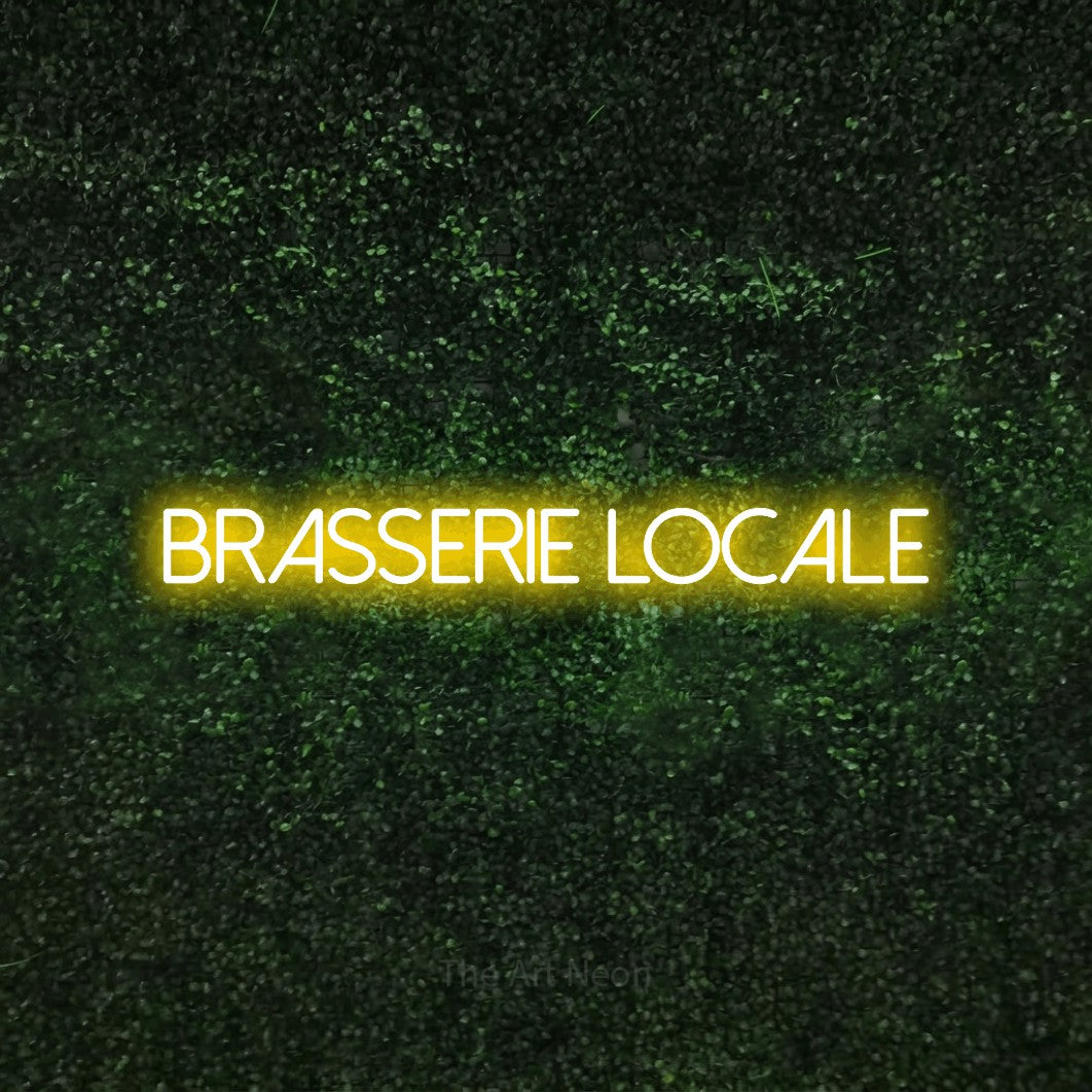 “Brasserie locale” LED neon sign in yellow, with thin sans-serif lettering, shown against a textured green hedge.