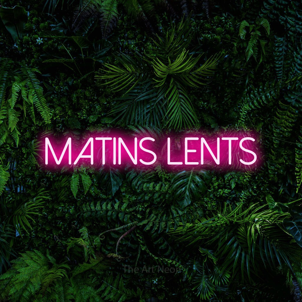 “Matins lents” LED neon sign in hot pink, with thin sans-serif lettering, shown against dense tropical foliage.