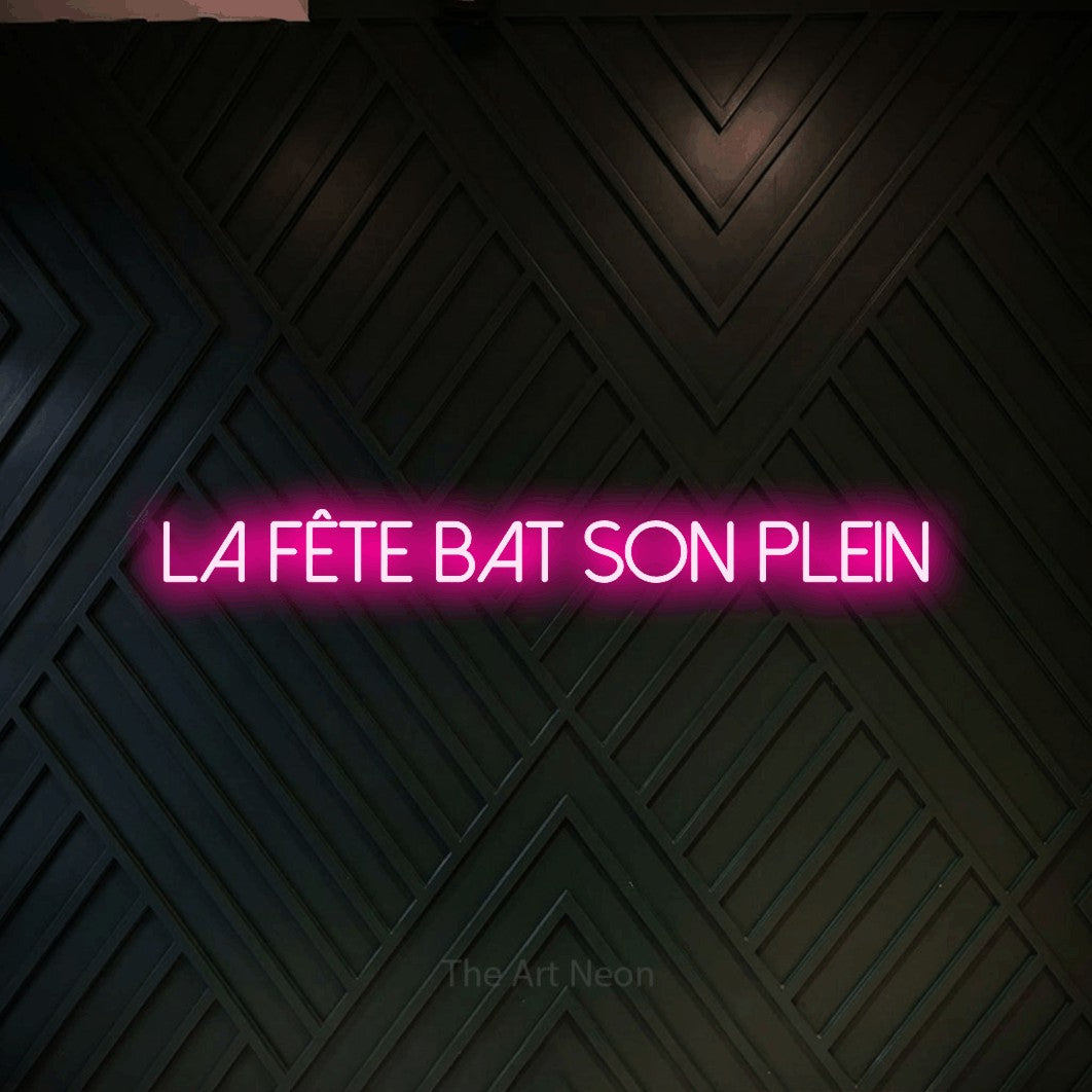 Composition en néon LED « La fête bat son plein » en rose vif, en lettres fines sans empattement, mise en valeur sur un mur en bois noir à motif chevron, pour une fête.