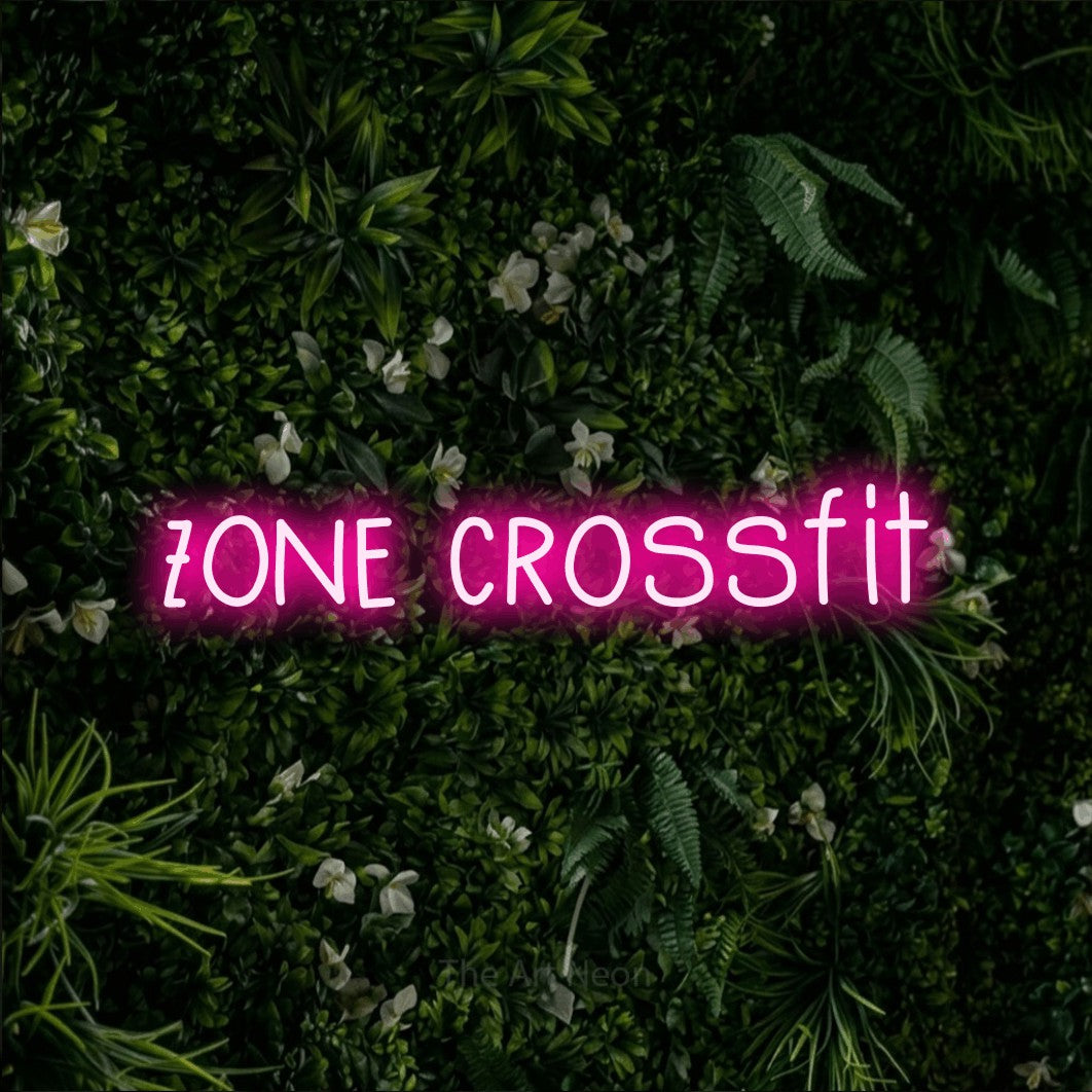 Lettrage en néon LED « Zone CrossFit » en rose vif, en lettres étroites dessinées à la main, présenté sur un mur végétal vert et dense, pour un espace d’entraînement.