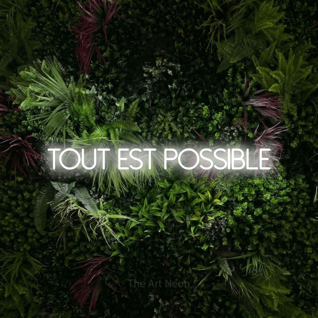 “Tout est possible” LED neon sign in cool white, with thin sans-serif lettering, shown against a plant wall with pink flowers.