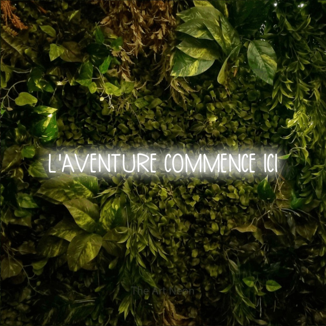 “L'aventure commence ici” LED neon sign in cool white, with narrow hand-drawn lettering, shown against bright green and yellow foliage.