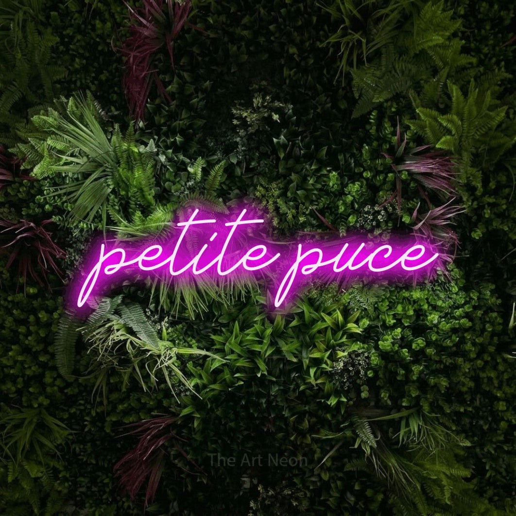 “Petite puce” LED neon sign in pink, with cursive lettering, shown against a plant wall with pink flowers.