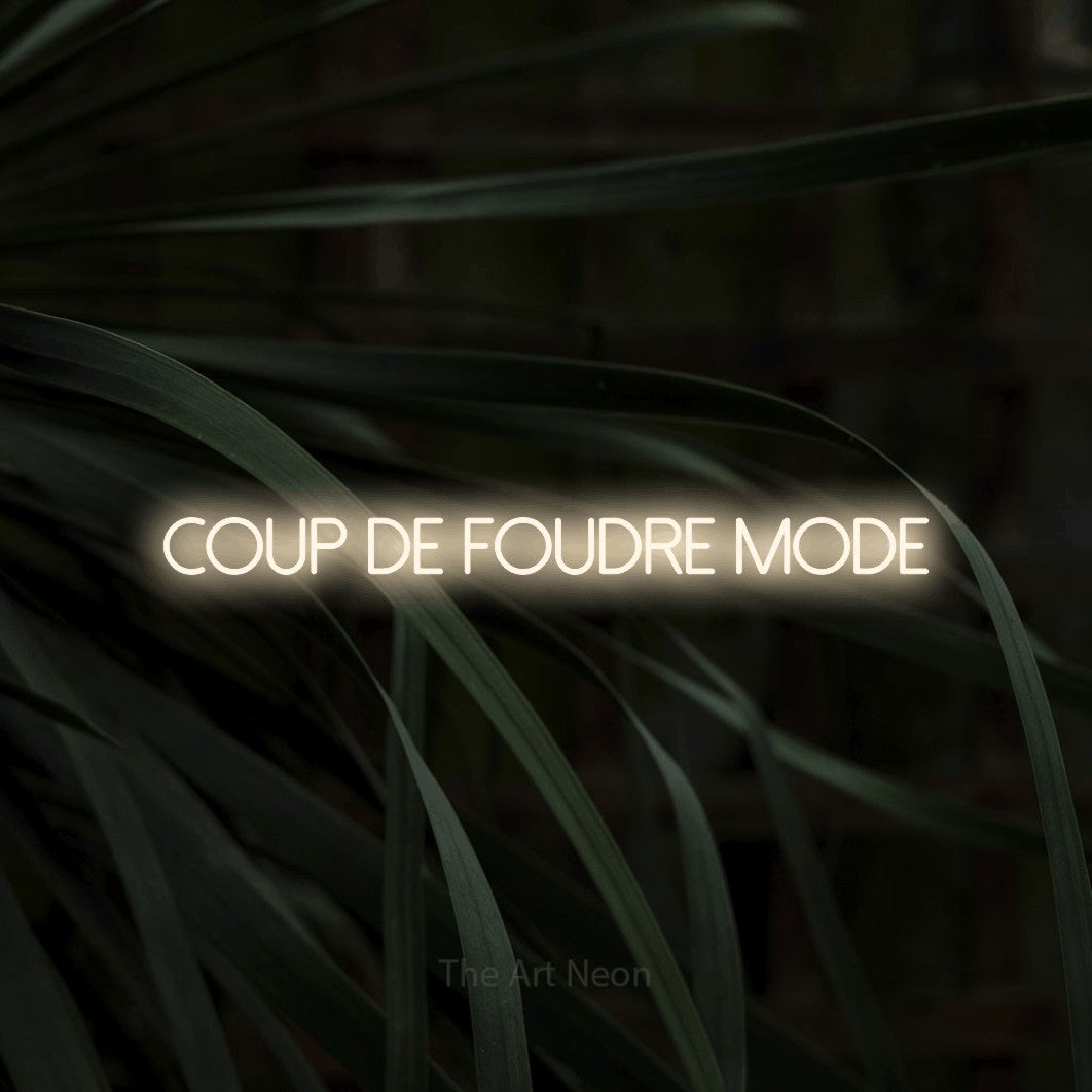 “Coup de foudre mode” LED neon sign in warm white, with thin sans-serif lettering, shown against a close-up of dark foliage.