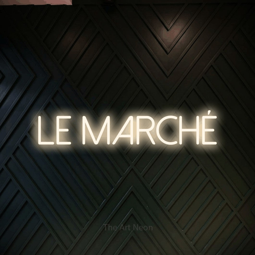 “Le marché” LED neon sign in warm white, with thin sans-serif lettering, shown against a black chevron-patterned wood wall.