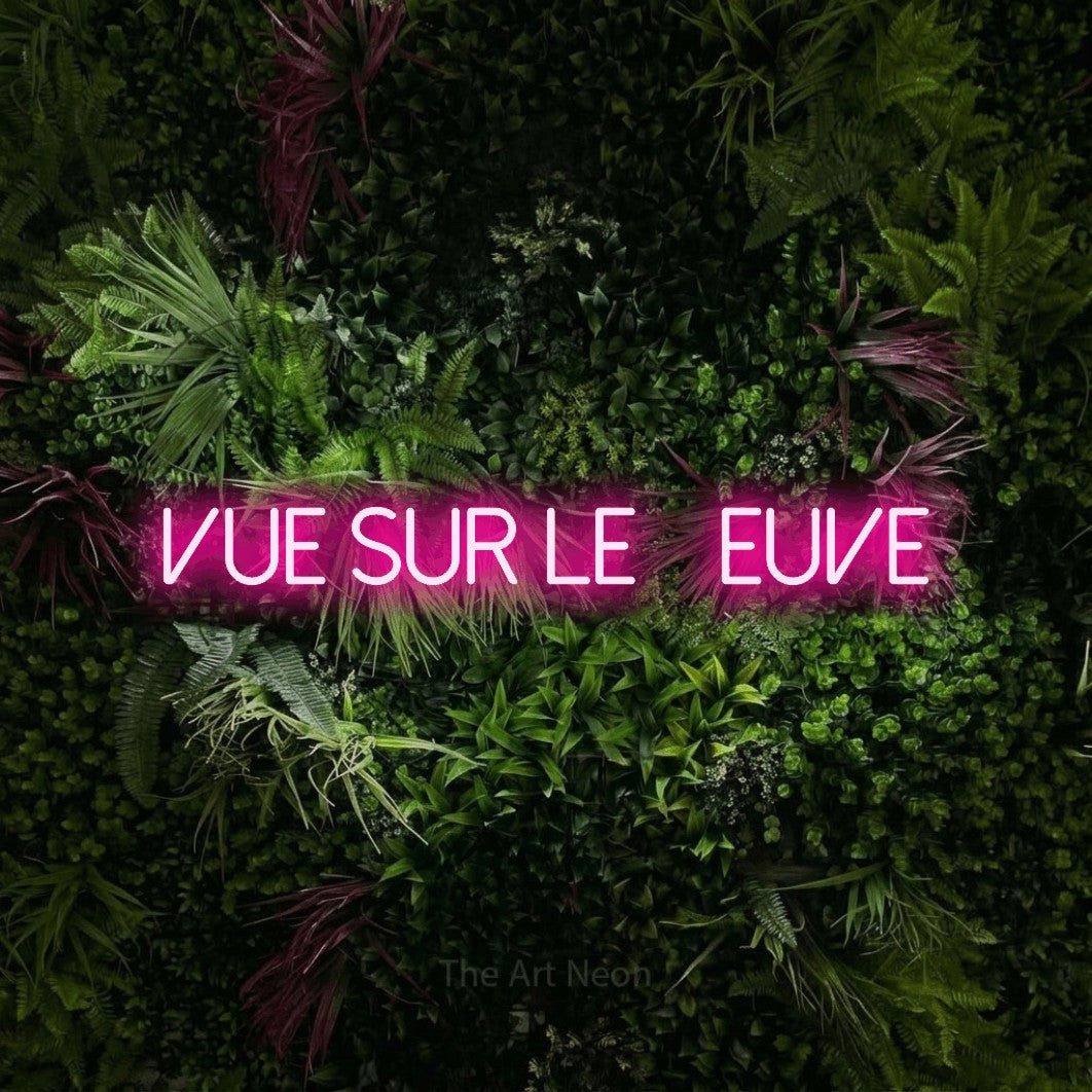 “Vue sur le fleuve” LED neon sign in hot pink, with thin sans-serif lettering, shown against a plant wall with pink flowers.