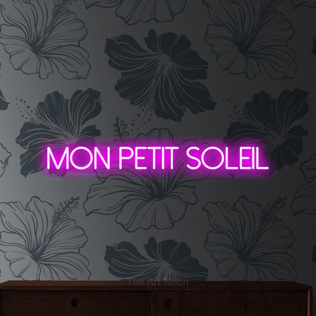 “Mon petit soleil” LED neon sign in pink, with thin sans-serif lettering, shown against a grey floral-patterned wall.