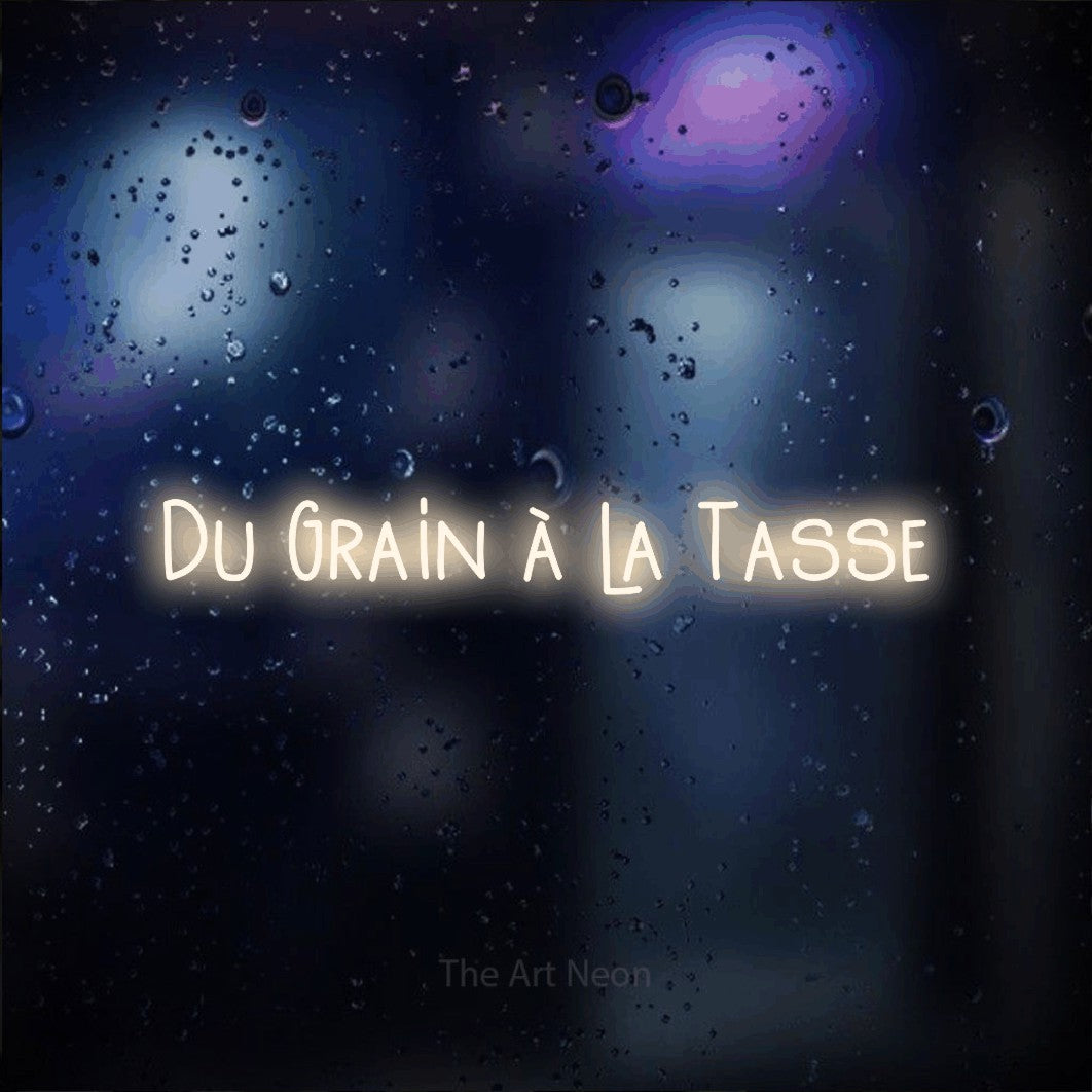 “Du grain à la tasse” LED neon sign in warm white, with narrow hand-drawn lettering, shown against glass with blue and purple reflections.