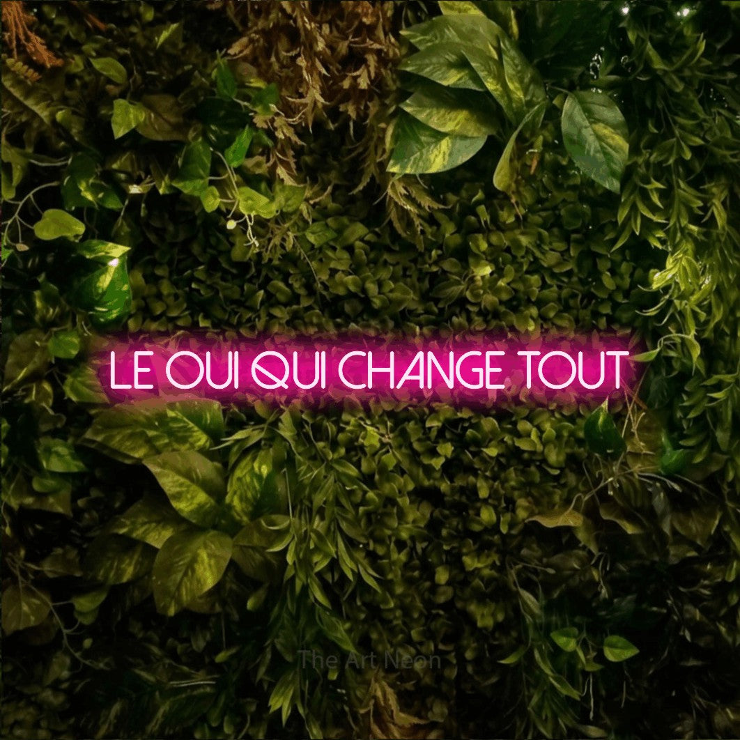 “Le oui qui change tout” LED neon sign in hot pink, with thin sans-serif lettering, shown against bright green and yellow foliage.