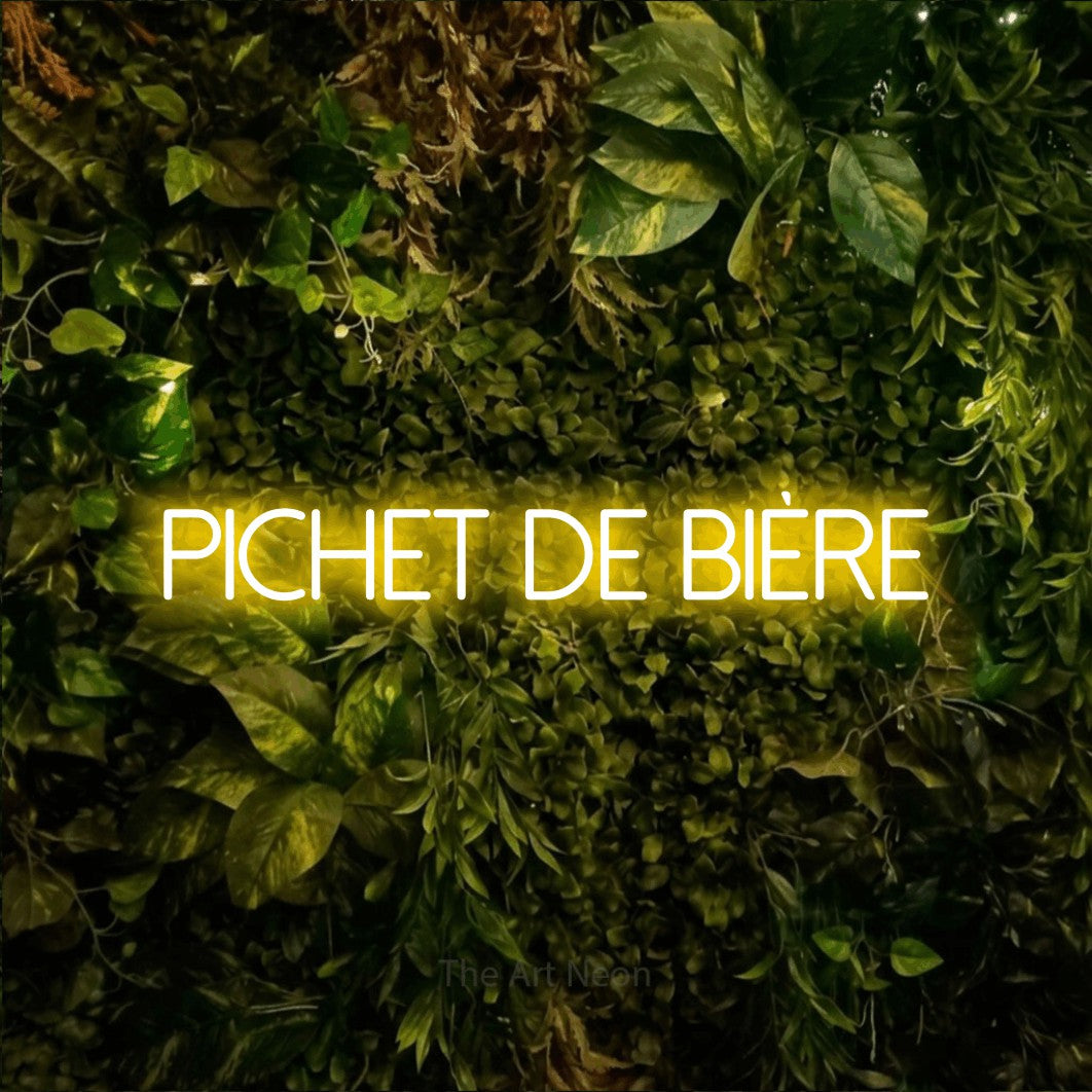 “Pichet de bière” LED neon sign in yellow, with thin sans-serif lettering, shown against bright green and yellow foliage.