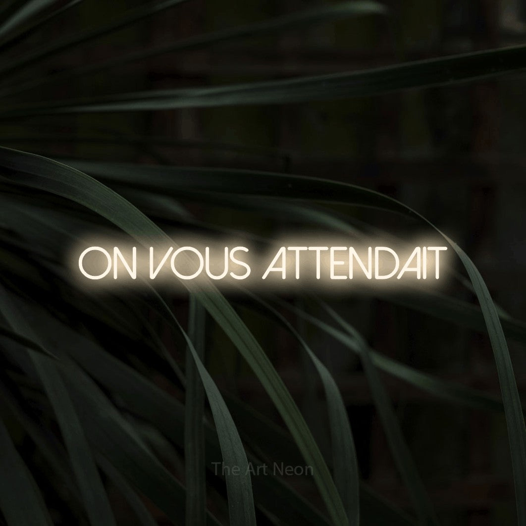 “On vous attendait” LED neon sign in warm white, with thin sans-serif lettering, shown against a close-up of dark foliage.