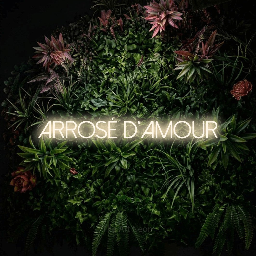 “Arrosé d'amour” LED neon sign in warm white, with thin sans-serif lettering, shown against a flowering plant wall.