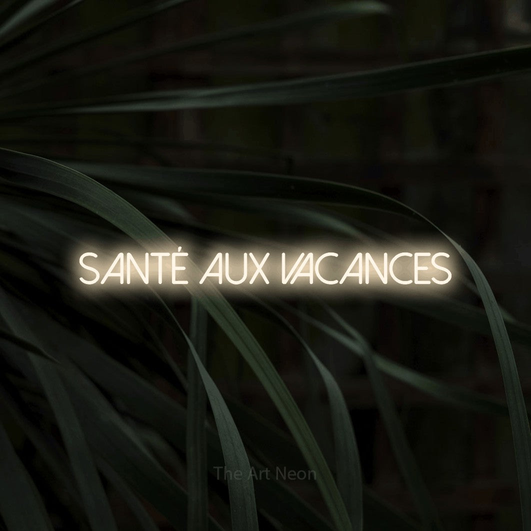 “Santé aux vacances” LED neon sign in warm white, with thin sans-serif lettering, shown against a close-up of dark foliage.
